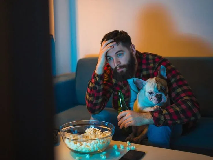 Frustrated viewer sitting with his dog in front of the TV, hand on his forehead, a bowl of popcorn on the table